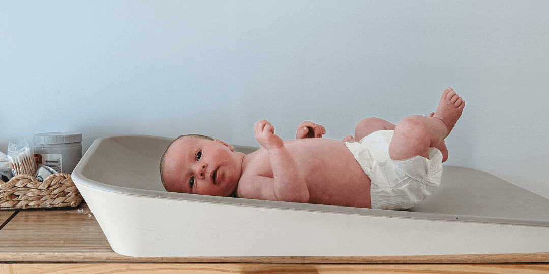 Newborn baby lying on a changing table wearing a nappy, with legs kicking up  - illustrating gentle, safe nappy options for newborns.