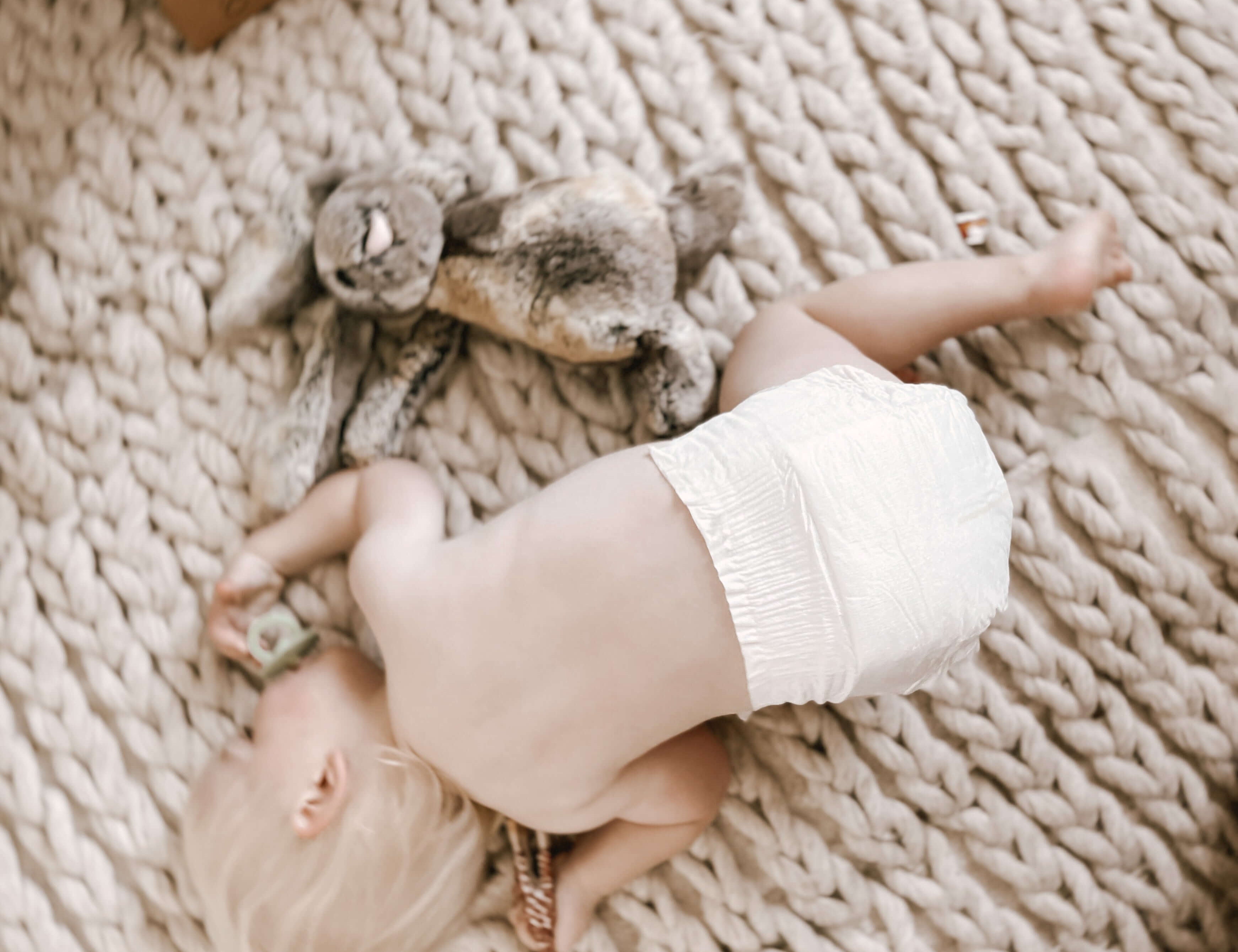 Baby wearing Cuddlies nappy lying on plus rug with toy